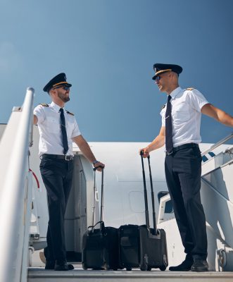 Full length portrait of handsome two men pilot in uniform with rolling bags in hands talking together in the outdoors